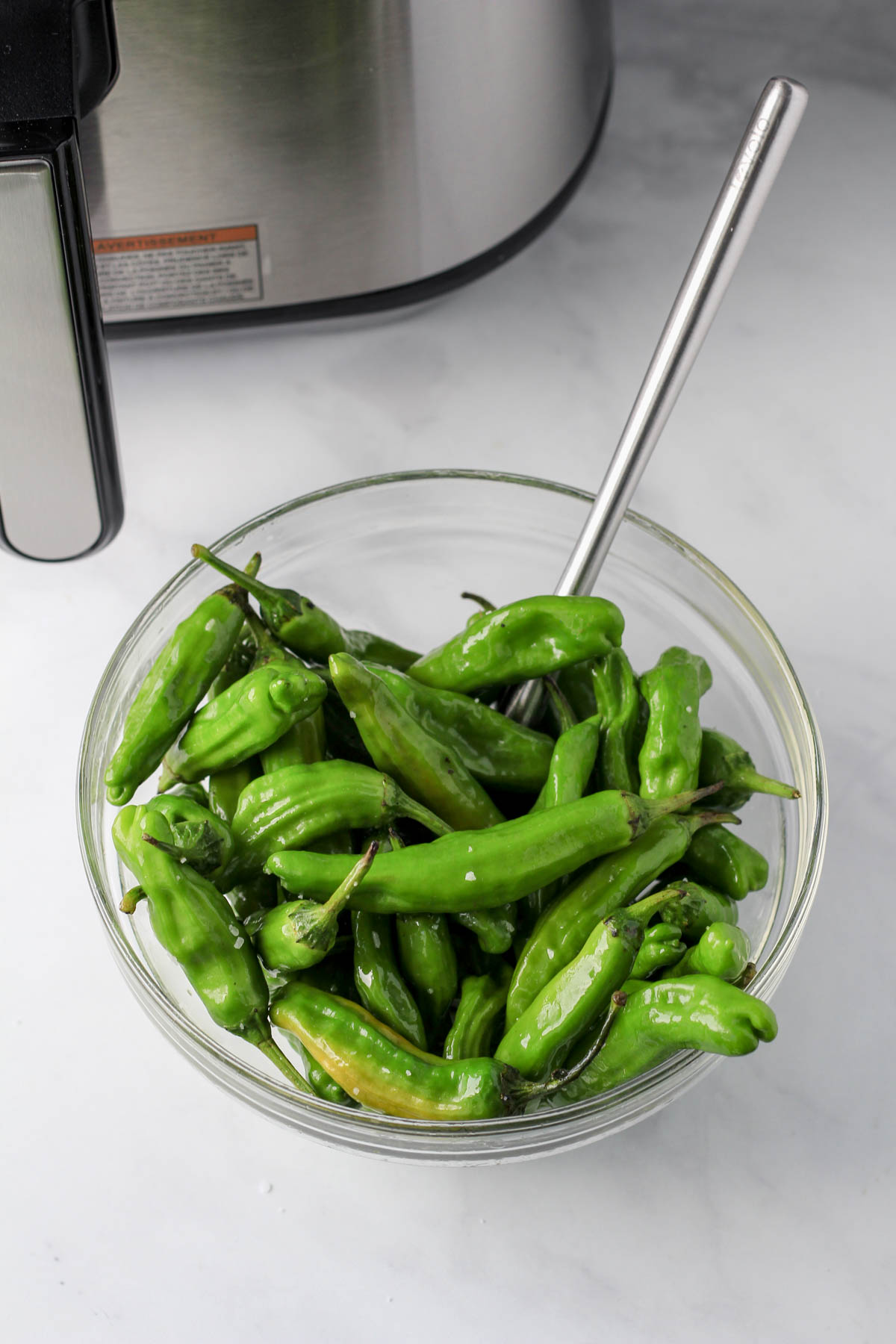 A small glass bowl with a spatula and shishito peppers coated in olive oil and salt.