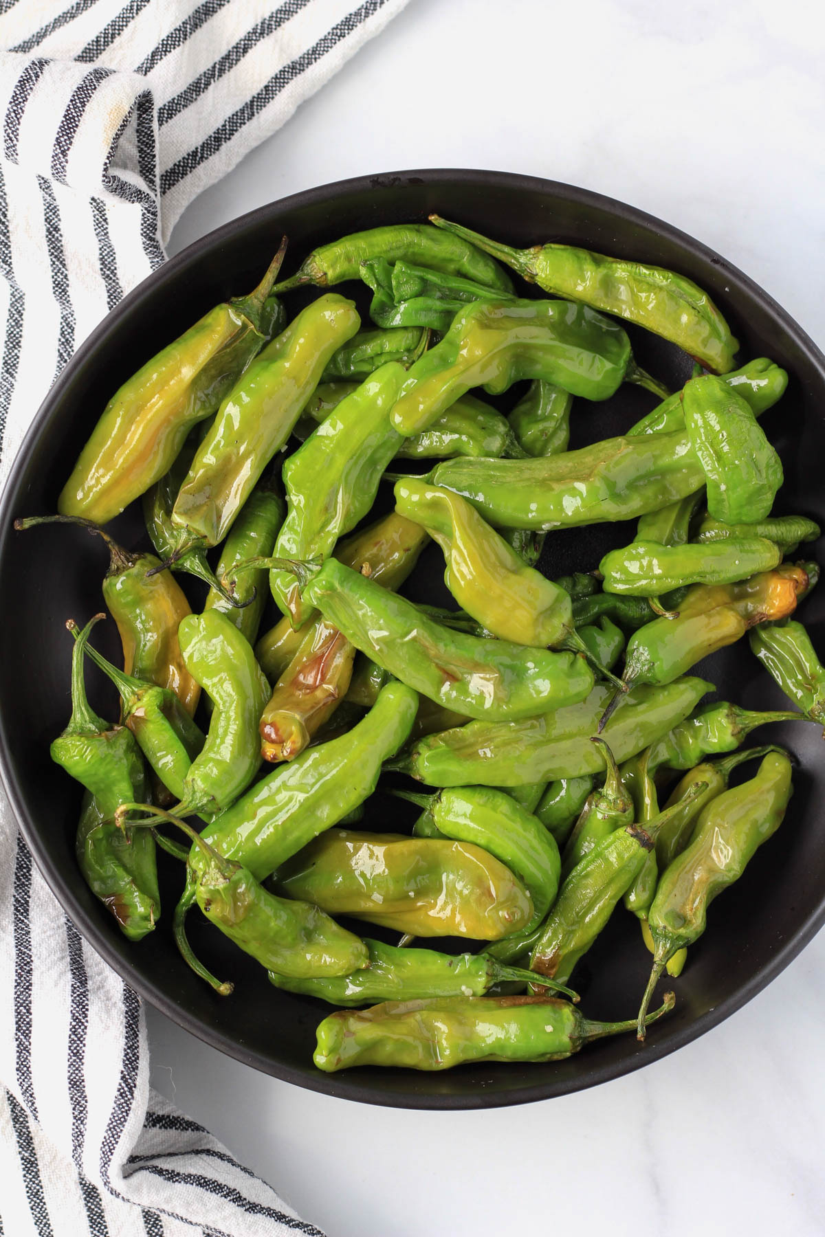 A black plate with cooked shishito peppers topped with salt.