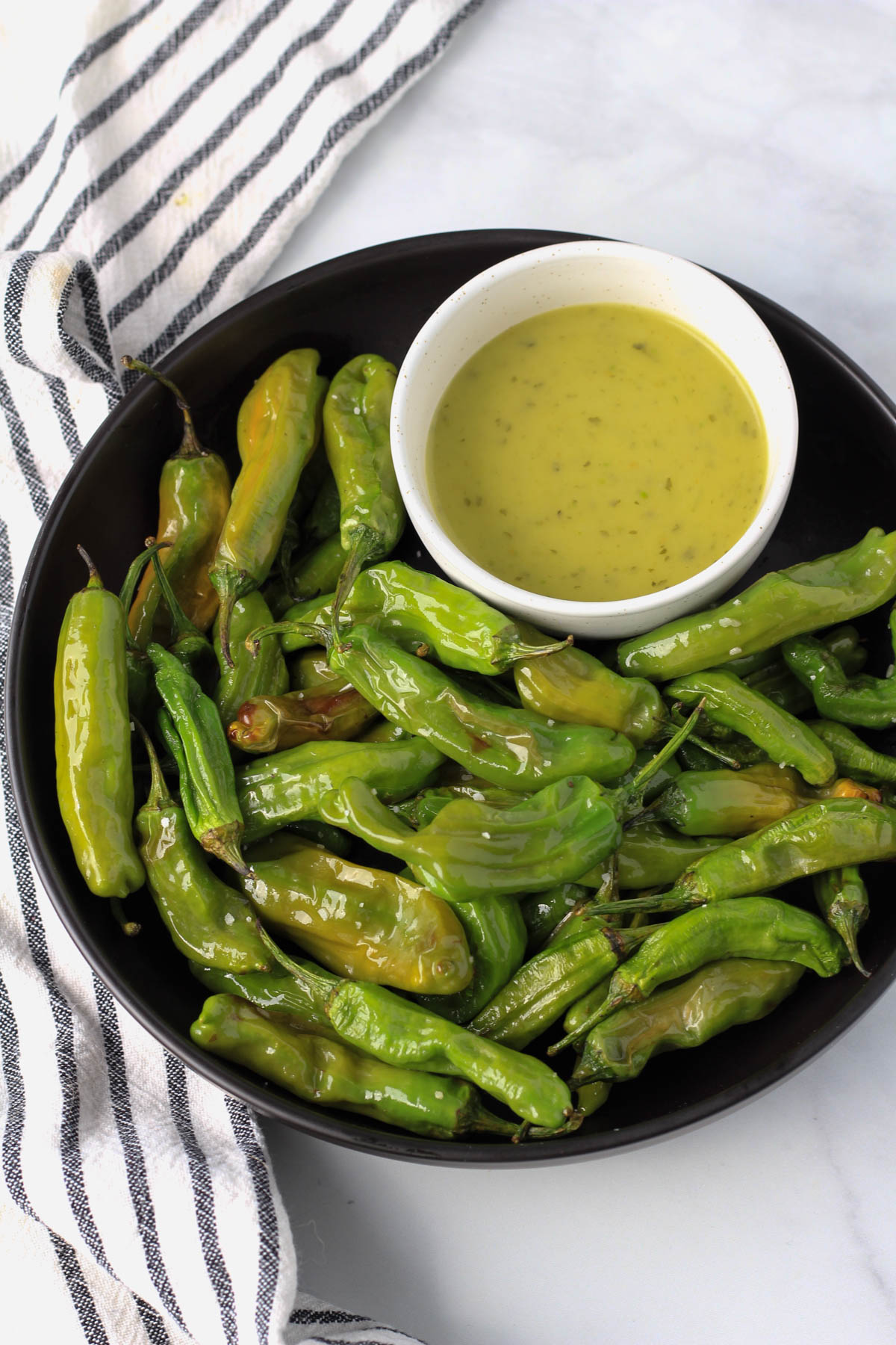 Blistered shishito peppers in a black plate with a white bowl of avocado dip.