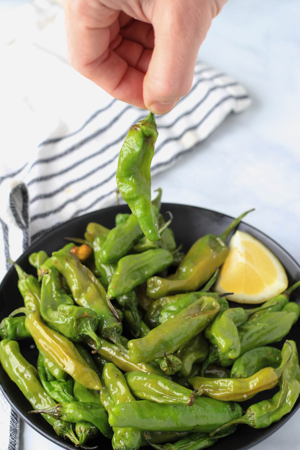 A hand picking up a shishito pepper off a black plate of blistered shishito peppers.