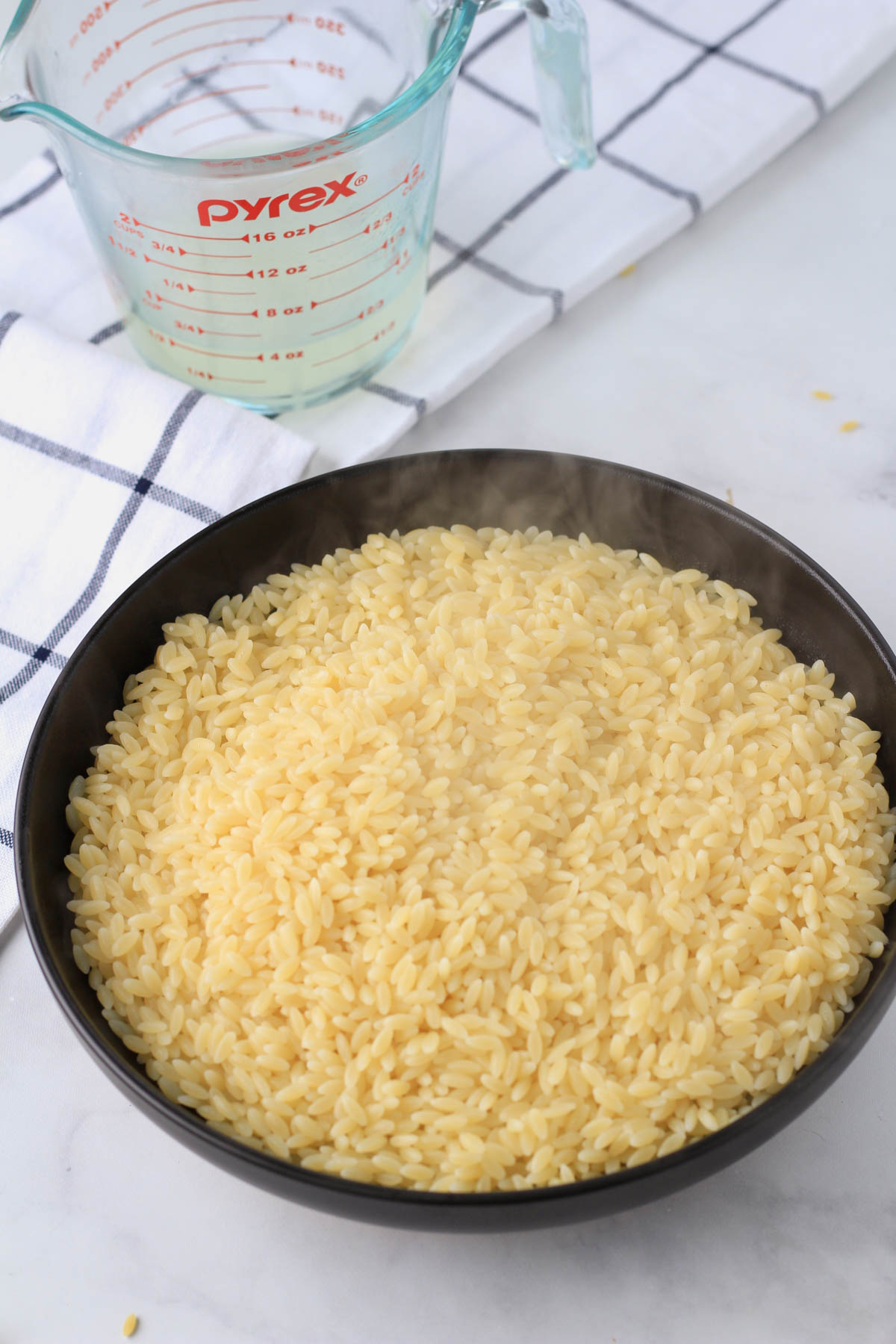 A black bowl with hot orzo pasta freshly cooked and a measuring cup in the back with reserved pasta water.