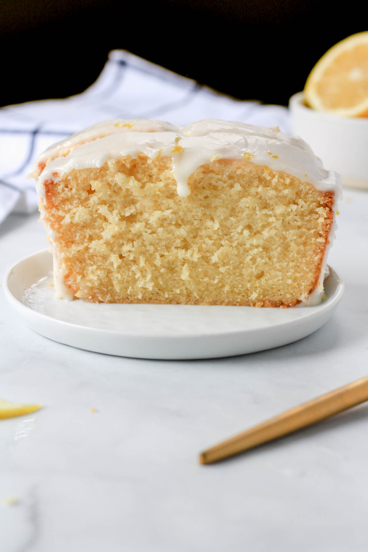 A slice of lemon loaf cake on a small white plate with a small bowl of lemon slices in the back right.