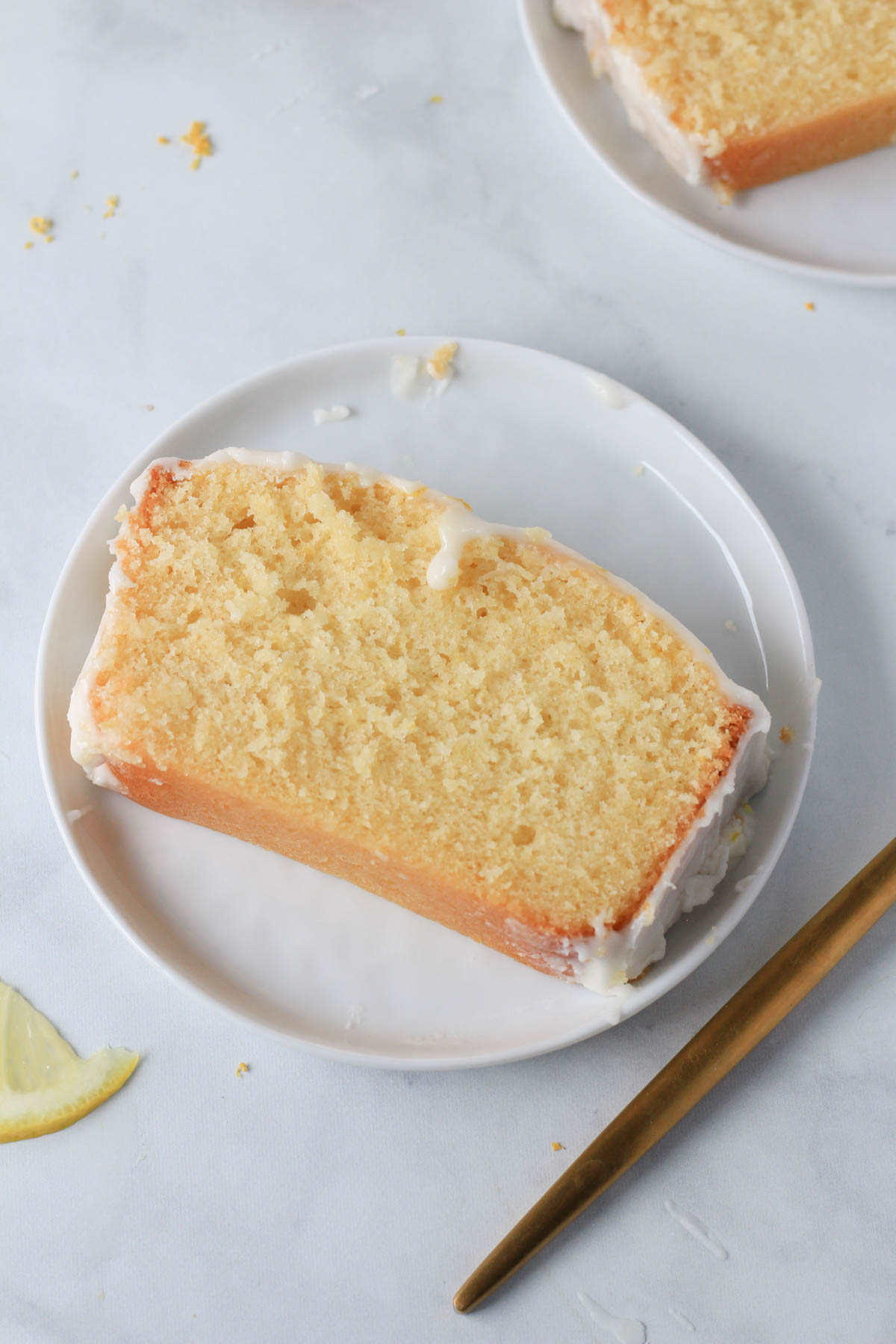 A top down picture of a slice of lemon loaf cake on a white plate with a gold fork to the right.