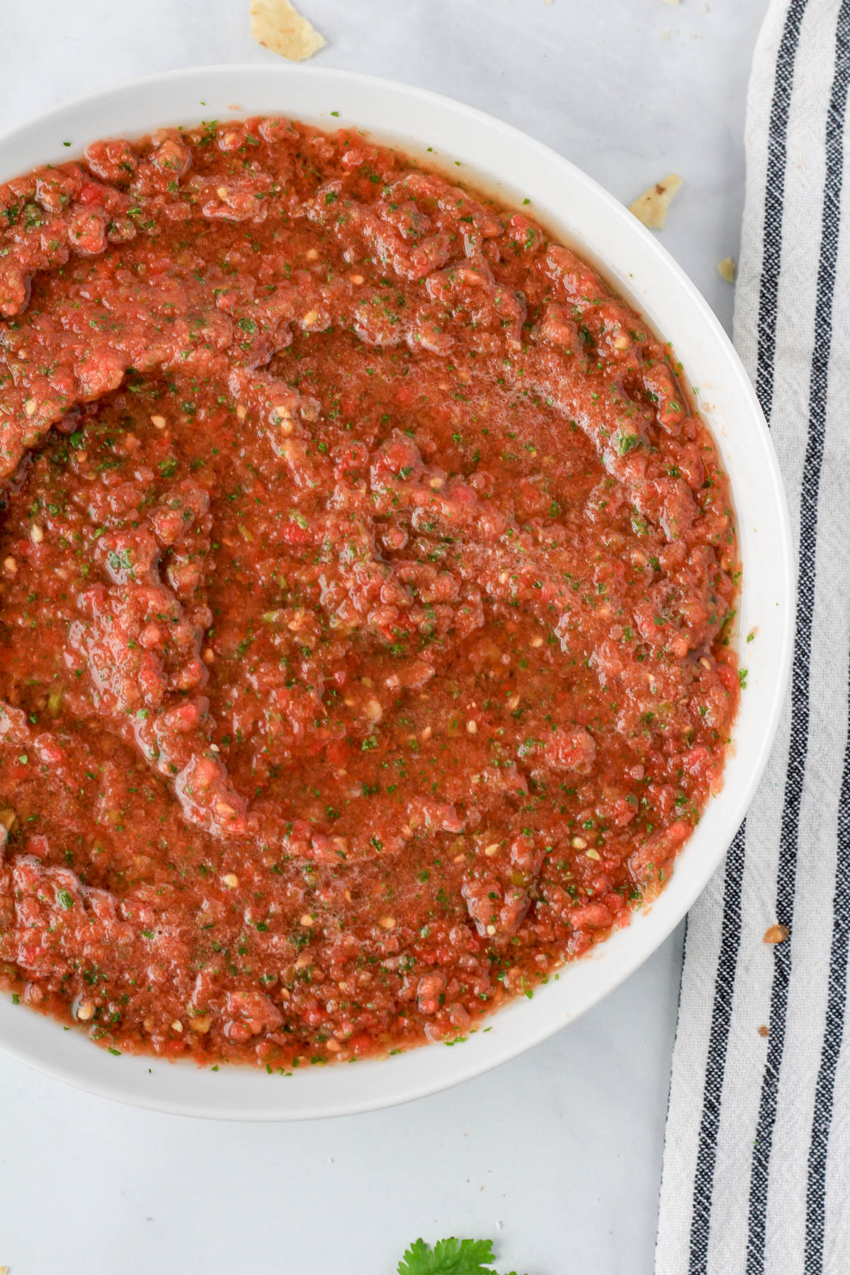 A close up of a white bowl of blender salsa on the left side of the image with a white and blue striped towel on the right side.