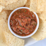 A close up of a small white bowl of blender salsa surrounded by tortilla chips.