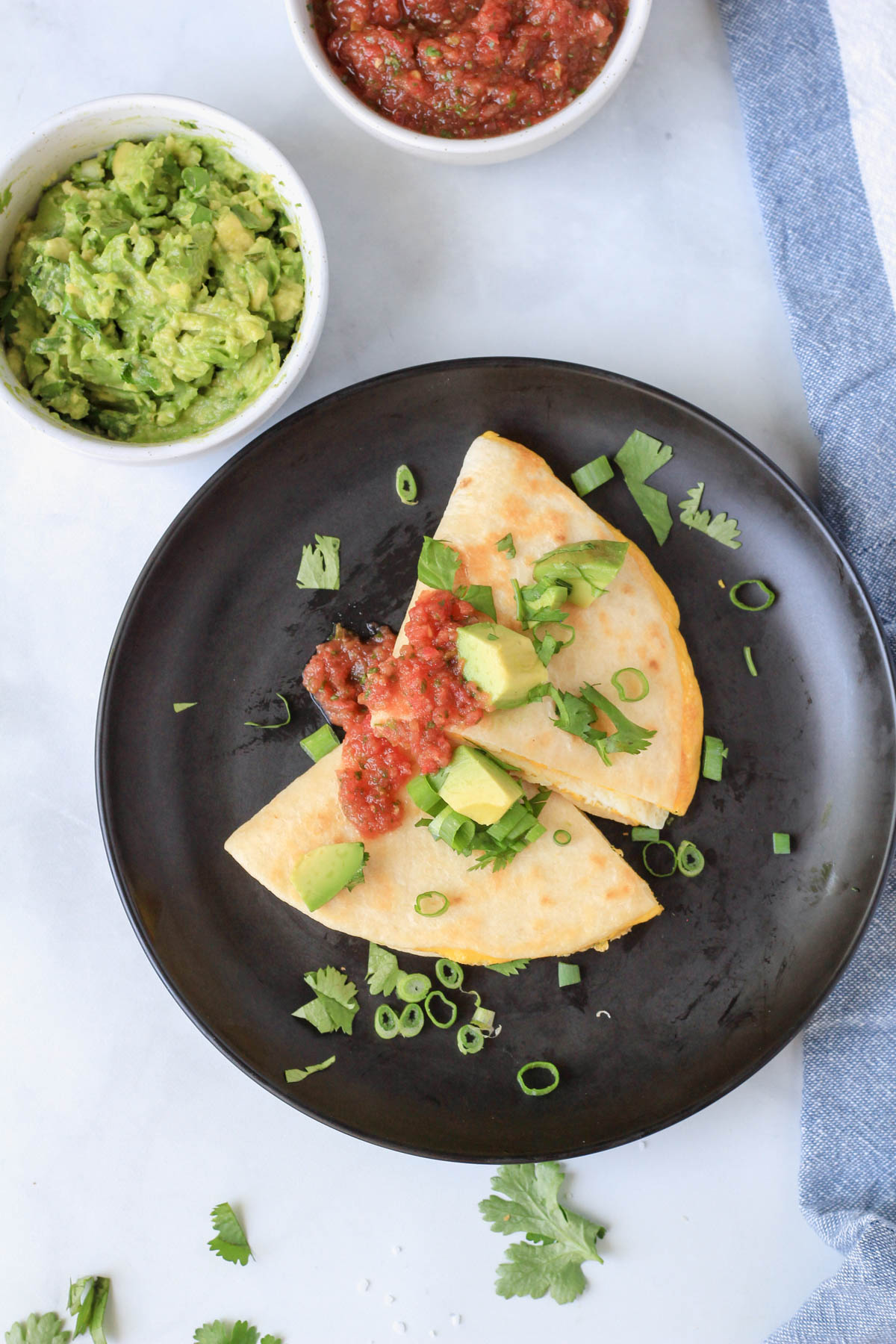 A top down photo of a black plate with two wedges of egg-a-dilla topped with salsa, green onion, and sliced avocado.