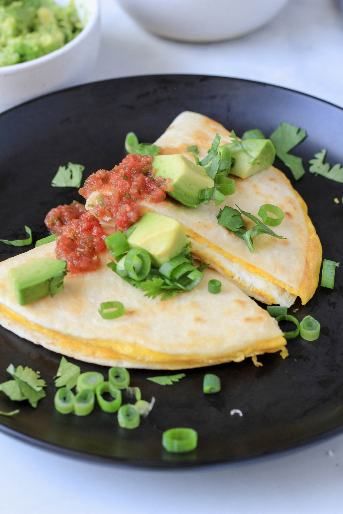 A black plate with two wedges of egg quesadilla topped with green onion, avocado, and homemade salsa.