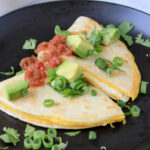 Two wedges of egg-a-dilla on a black plate topped with green onion, avocado, and salsa.