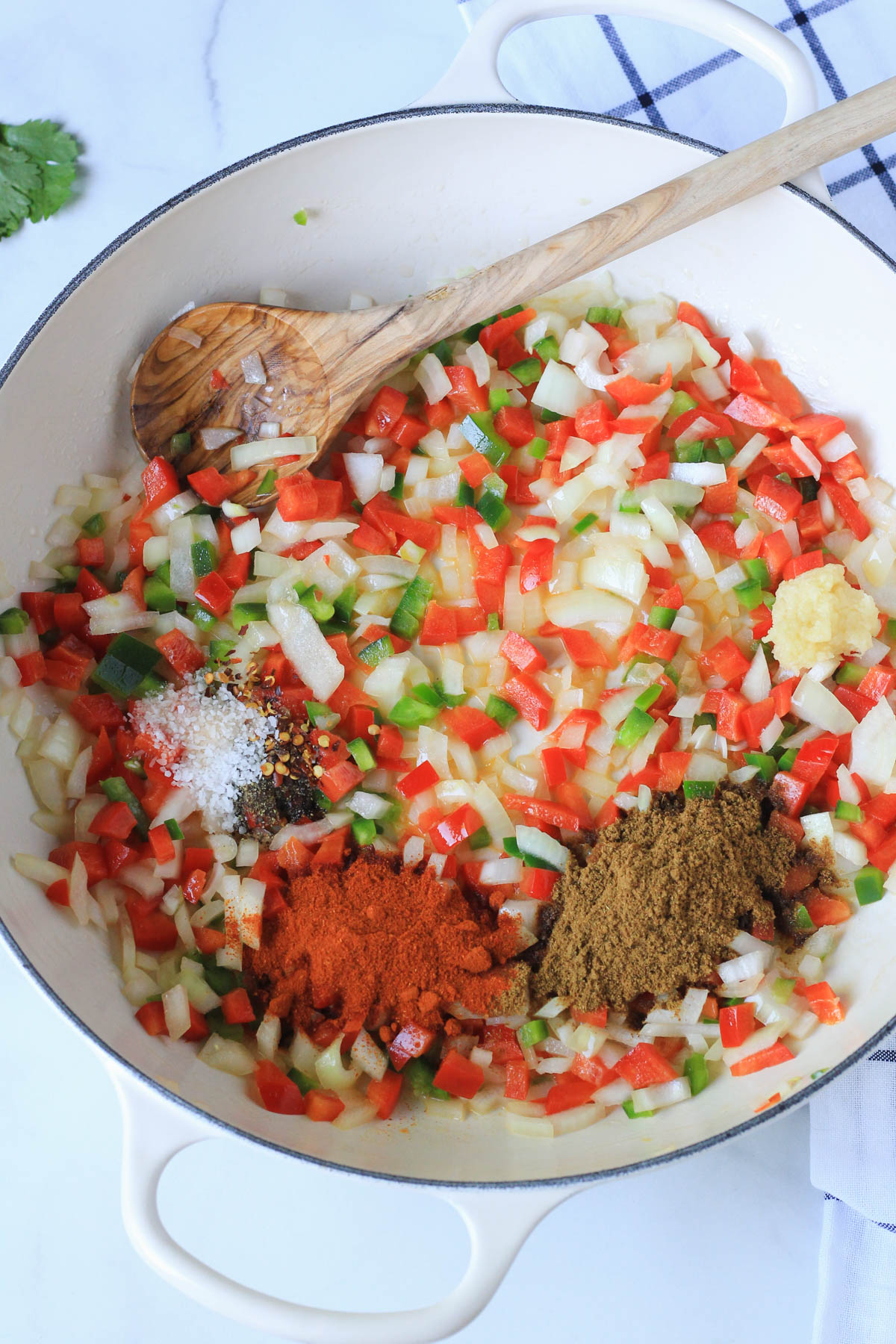 A white skillet with a wooden spoon and vegetables with spices cooking.