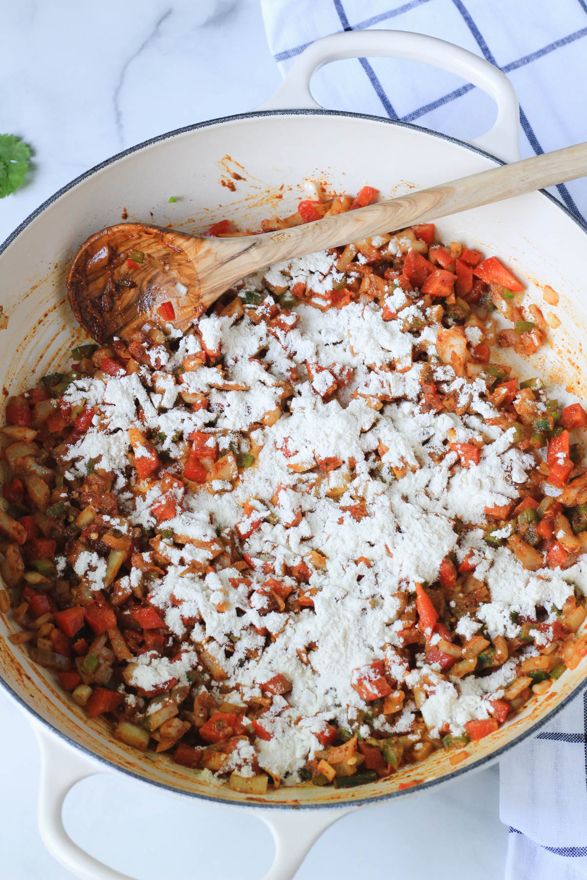 Flour sprinkled on top of the cooked vegetables in a white skillet with a wooden spoon.
