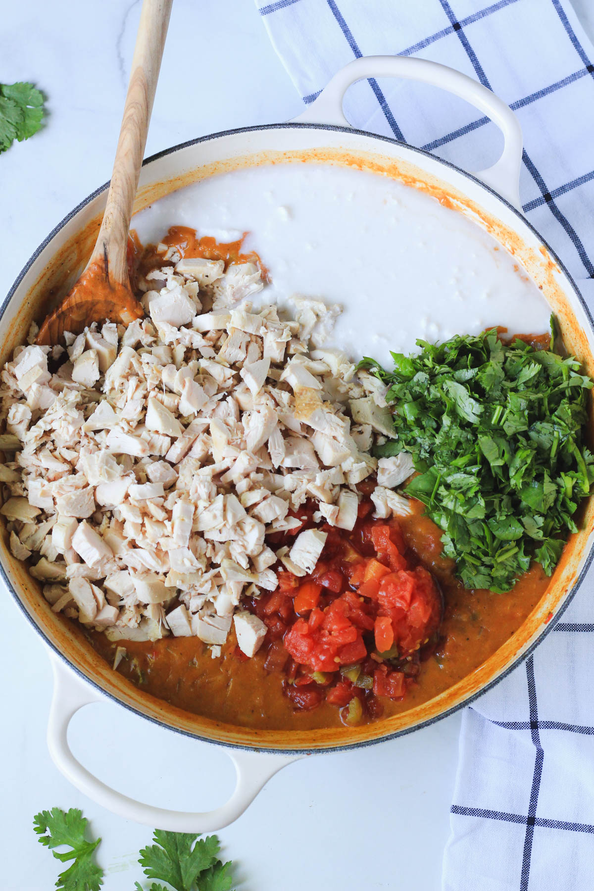 Chicken, rotel, coconut milk, and cilantro on top of the creamy king ranch filling in a white skillet with a wooden spoon on a white counter.