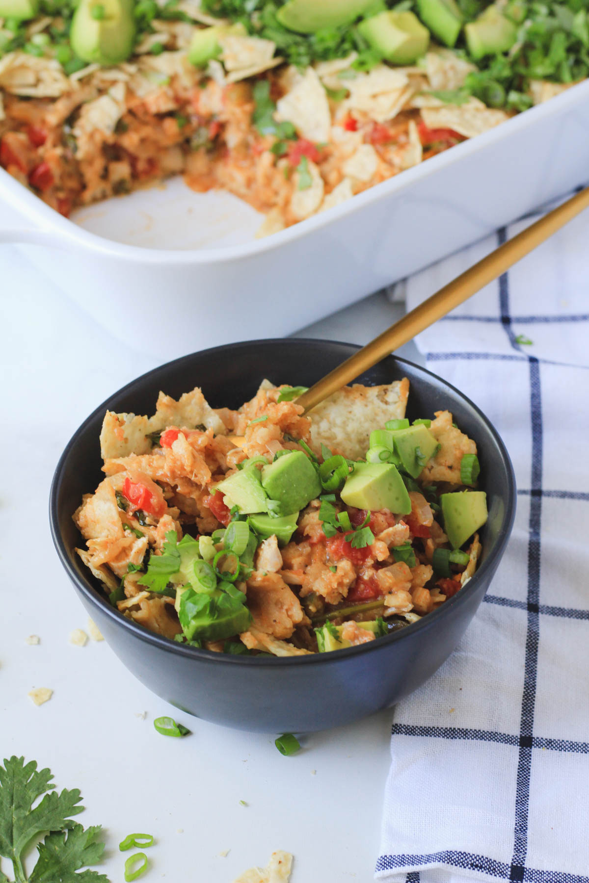 A dark blue bowl with a serving of king ranch chicken casserole topped with avocado, tortilla chips, and green onion with a gold fork in front of a white casserole pan of king ranch.
