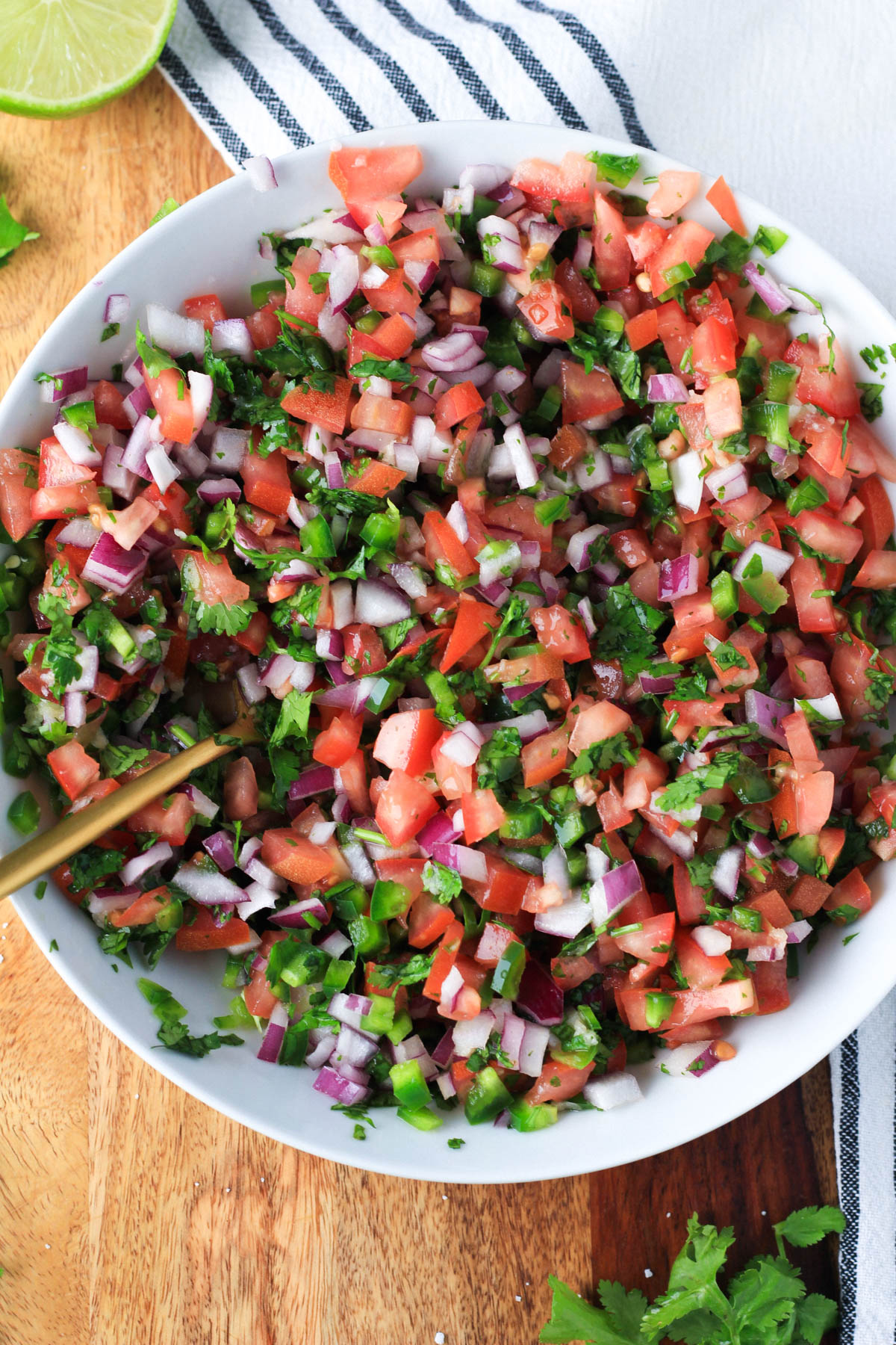 A white bowl with a gold spoon mixing the pico de gallo on a wooden counter.