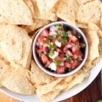 A white bowl filled with pico de gallo surrounded by tortilla chips in a white bowl.