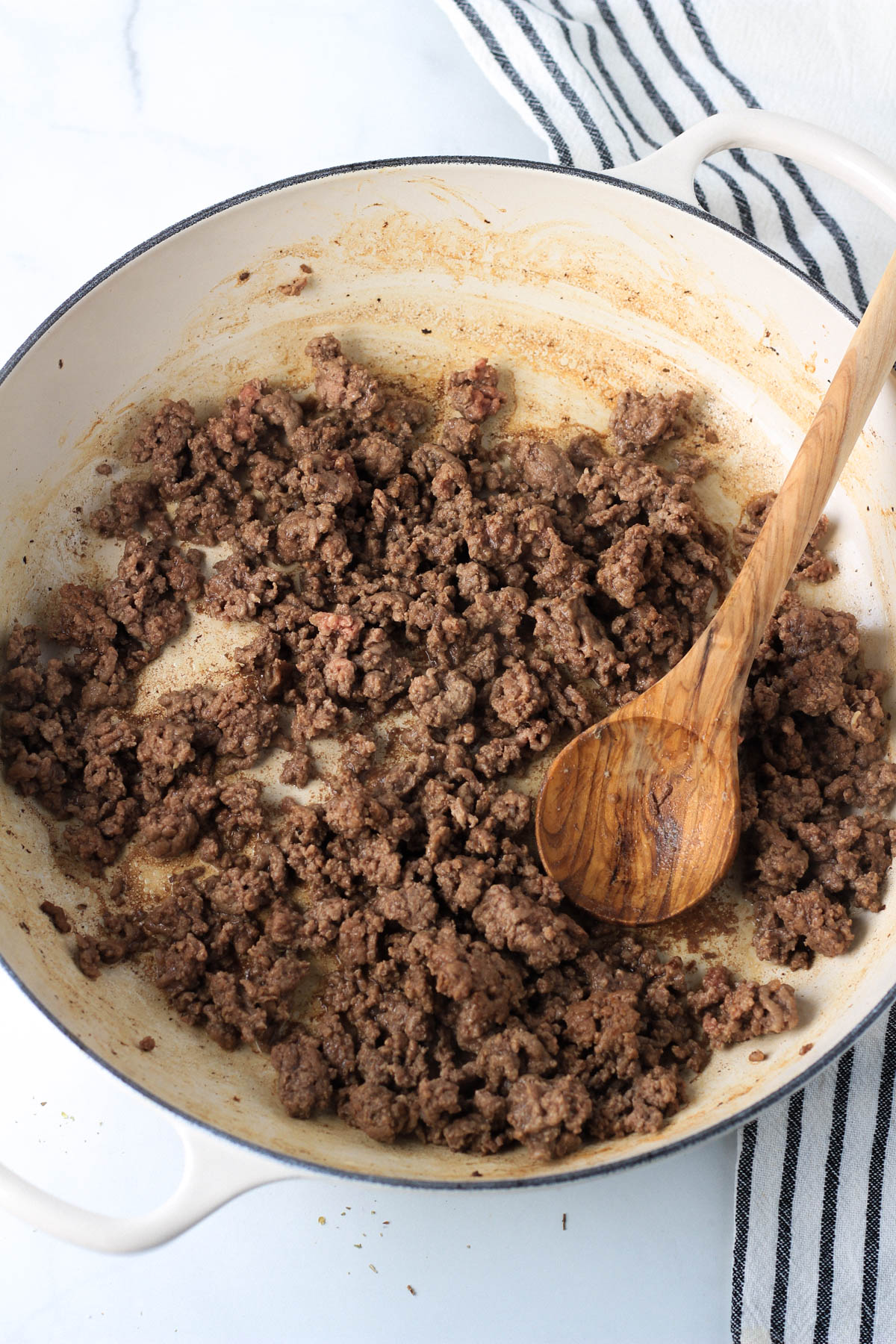 A large white skillet with a wooden spoon and browned ground beef.