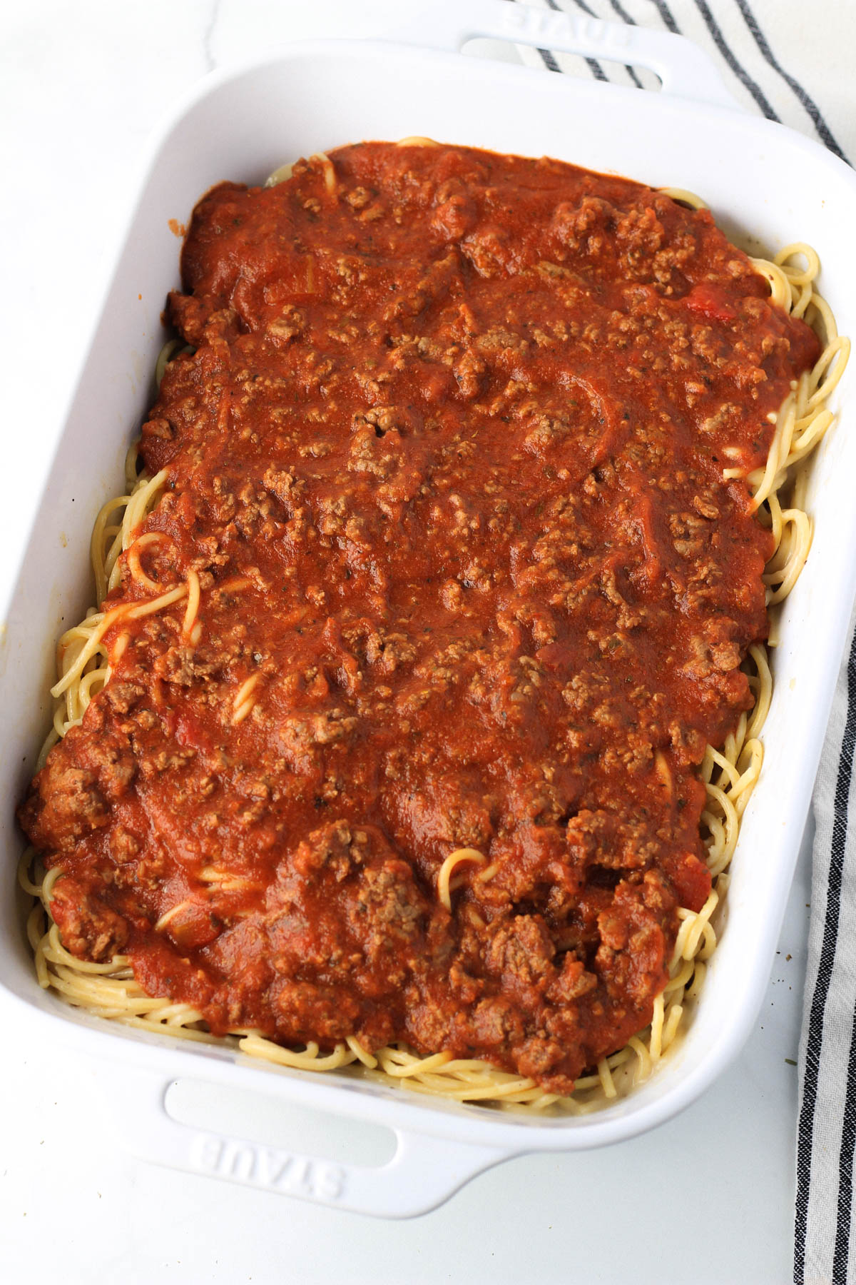 A large white baking dish with pasta coated in garlic cream sauce and topped with a meat sauce.