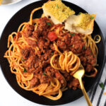 A small black plate with red and white pasta and a gold fork with a slice of garlic bread on the plate.