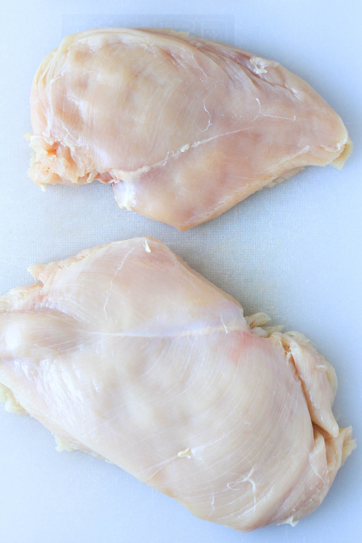 A white cutting board with two boneless skinless chicken breasts after being pounded flat.