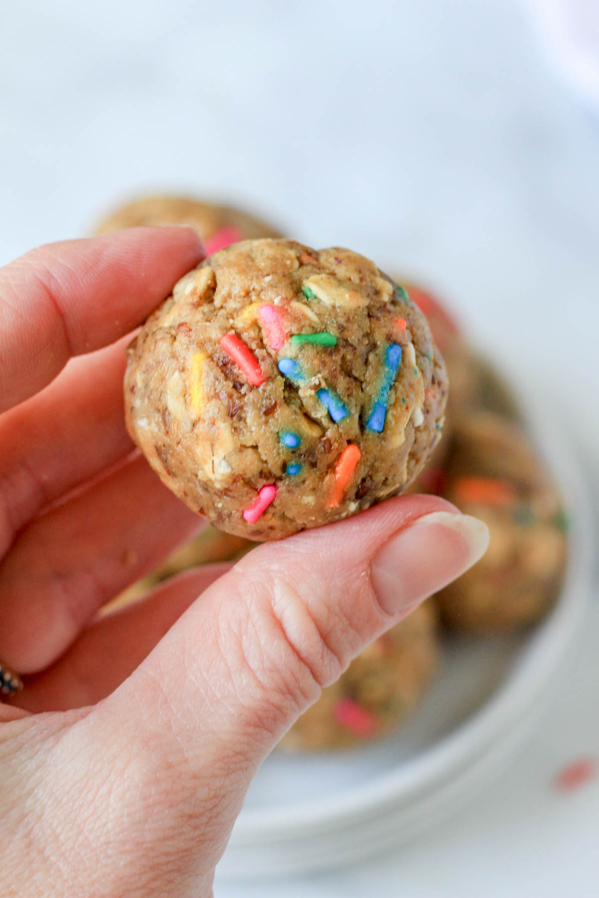 A hand holding one cake batter lactation bite in front of a plate filled with lactation bites.
