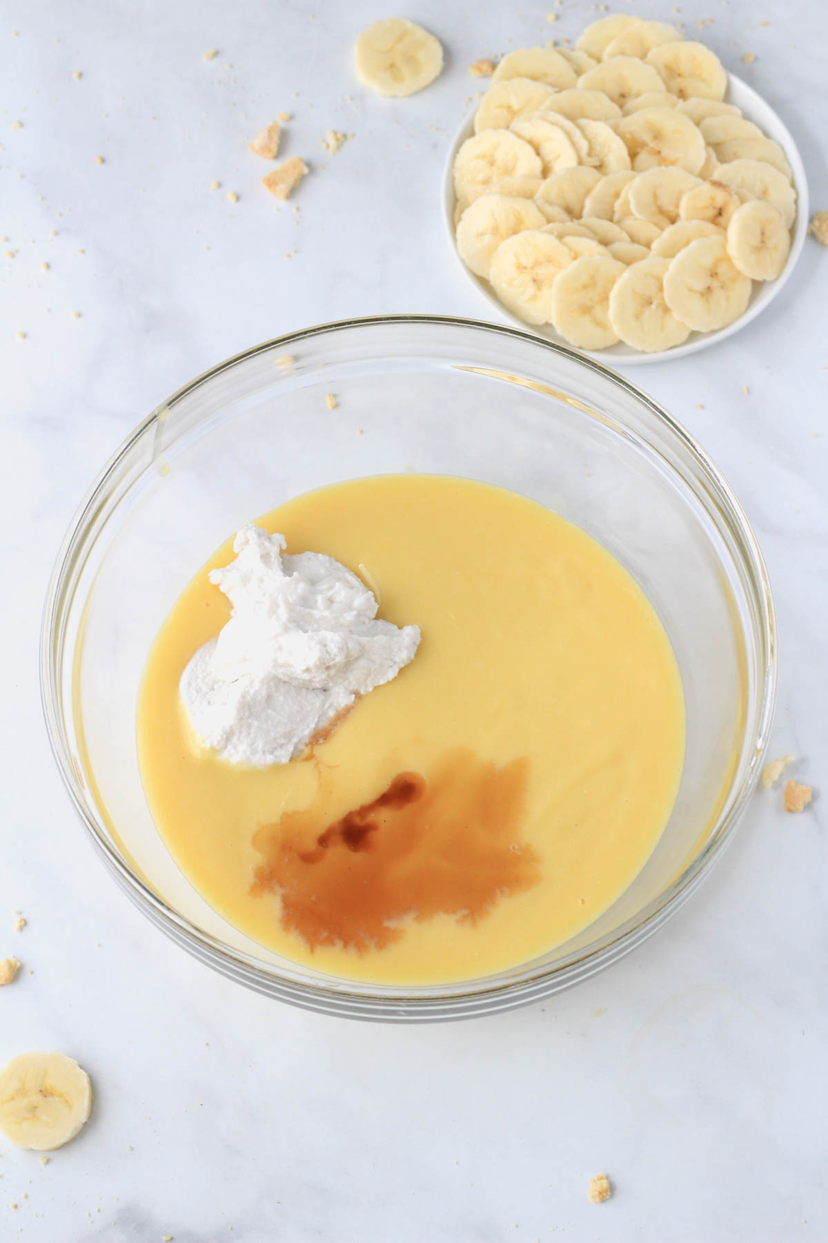 A glass bowl with banana pudding, coconut cream, and vanilla with a small plate of sliced banana in the top right corner.