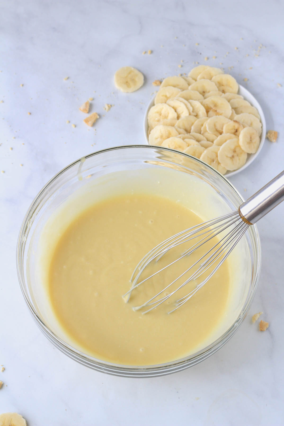 A glass bowl with banana pudding popsicle filling and a wire whisk with a small plate of sliced bananas in the top right.
