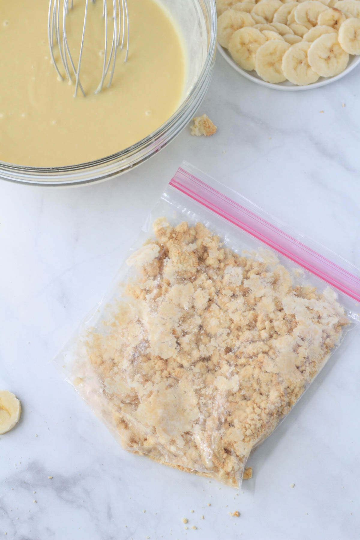 A ziplock bag with crushed golden oreos with a bowl of banana pudding filling in the top left.