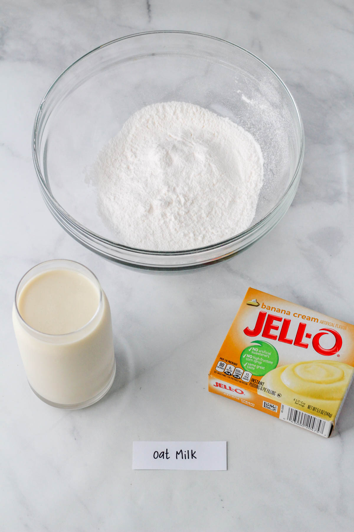 A glass of oat milk with a box of banana cream jell-o mix and a glass bowl.