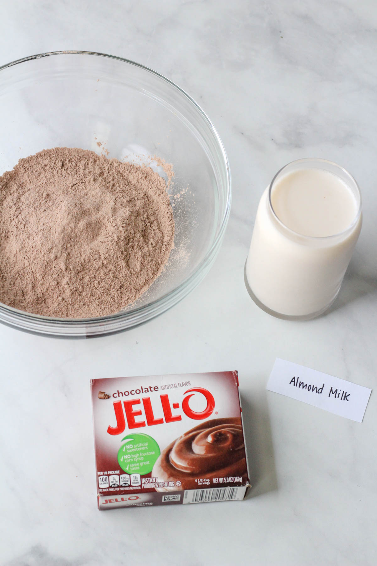 A glass bowl with chocolate jell-o pudding mix and a glass of almond milk to the right with the chocolate jell-o pudding box below.