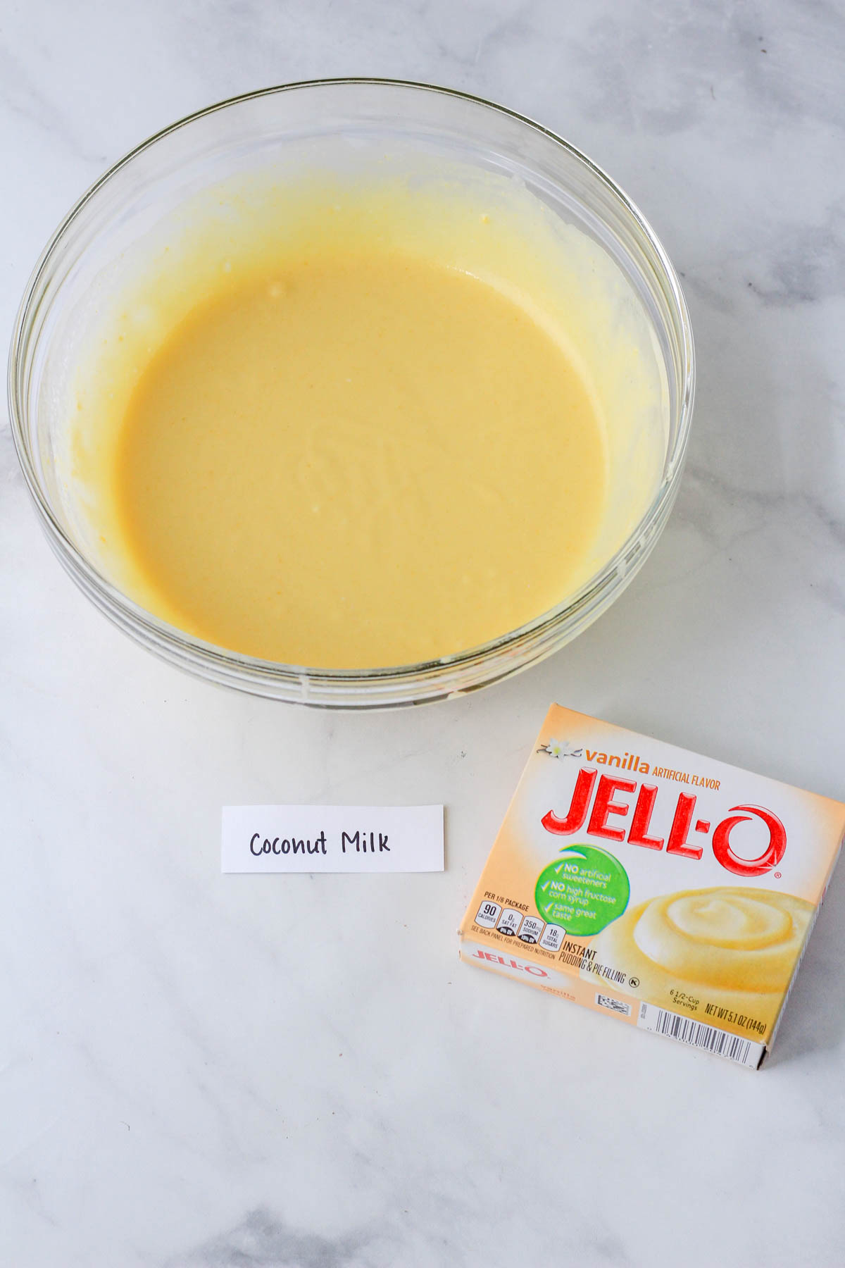 A glass bowl with vanilla pudding mixed with coconut milk and a box of vanilla jell-o to the right.