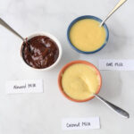 Three bowls with spoons and labels of instant pudding in a triangle on the top right a blue bowl with oat milk based jell-o, in the bottom right an orange bowl with coconut milk based jell-o, and on the right side a white bowl with almond milk based pudding.