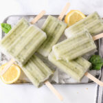 A silver cookie sheet with ice and lemon basil popsicles scattered on top.