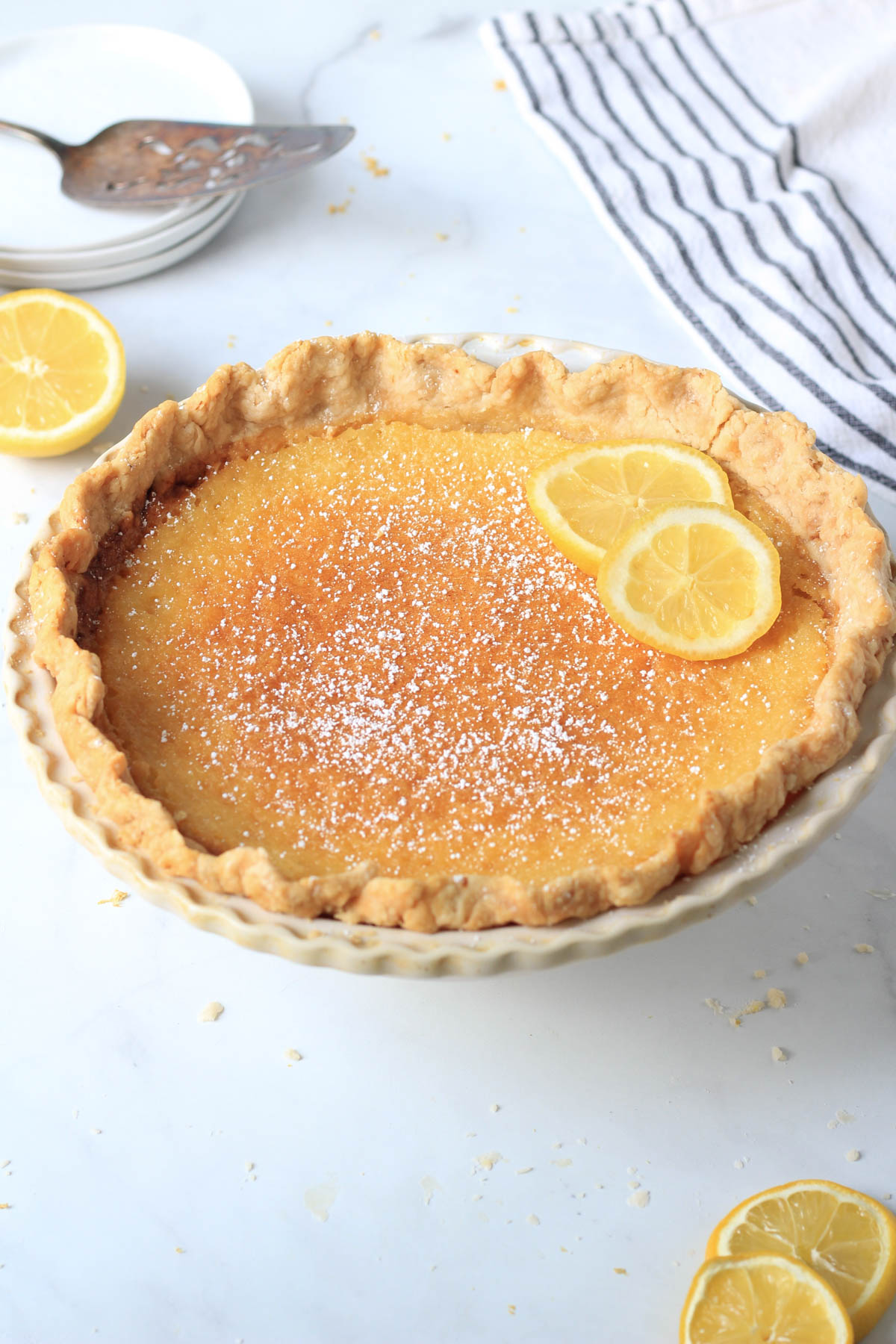 A lemon chess pie on a white counter with lemon slices and a pie server in the back left.