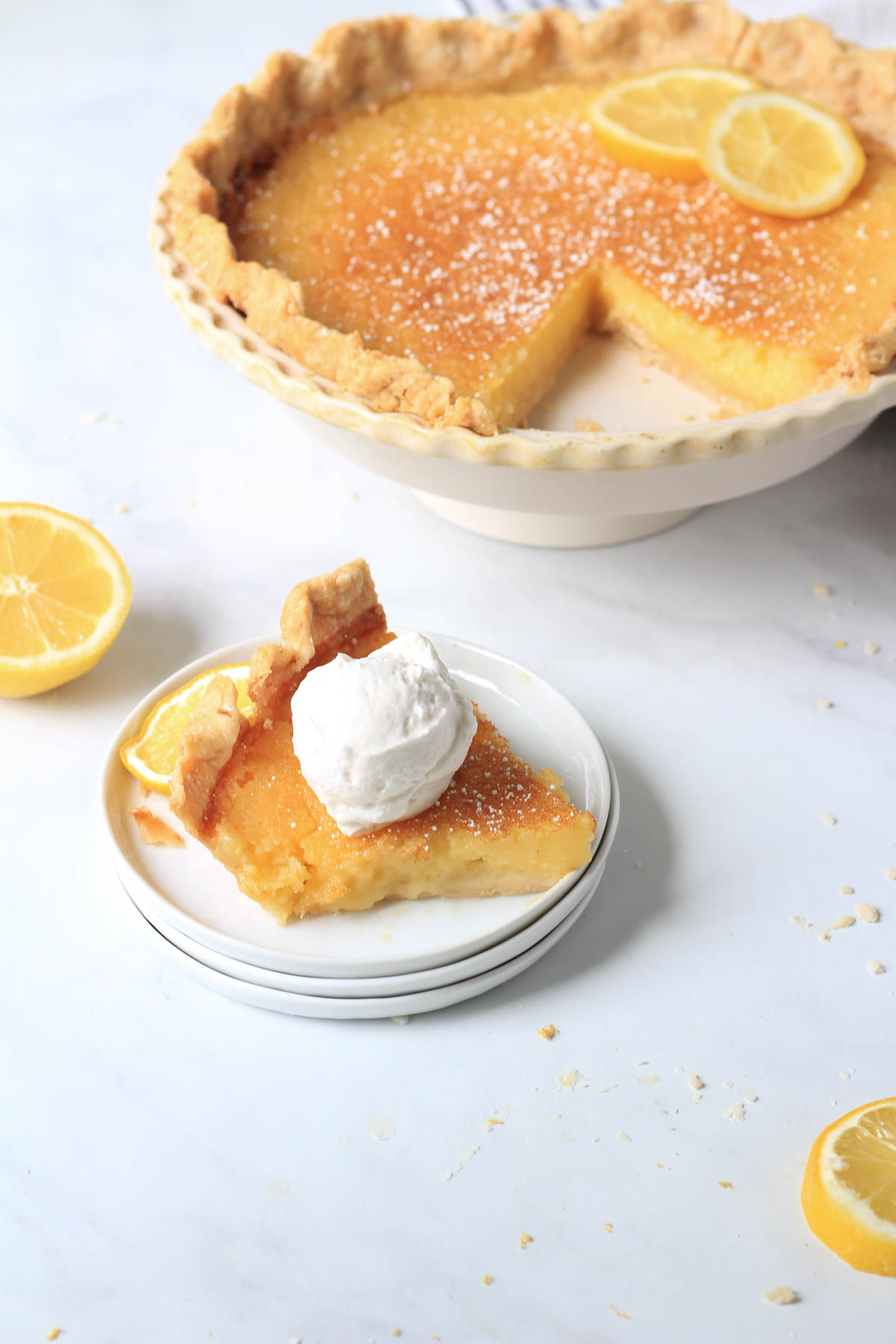 A small stack of white plates with a slice of dairy-free lemon pie topped with whipped topping and a pie pan of the remaining pie in the back right.