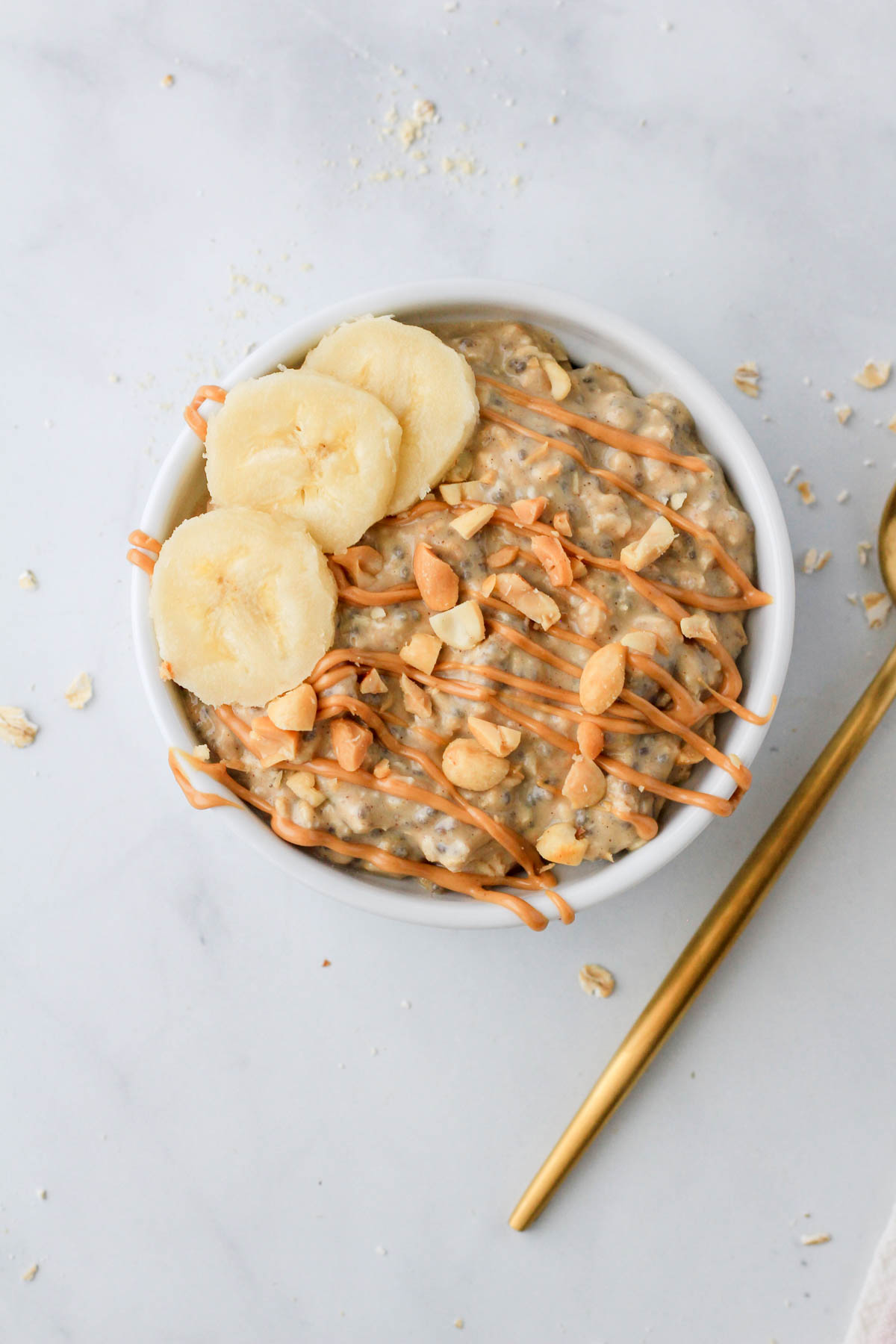 A top down picture of vegan vanilla overnight oats topped with peanut butter, peanuts, and banana slices in a white bowl on a white counter.