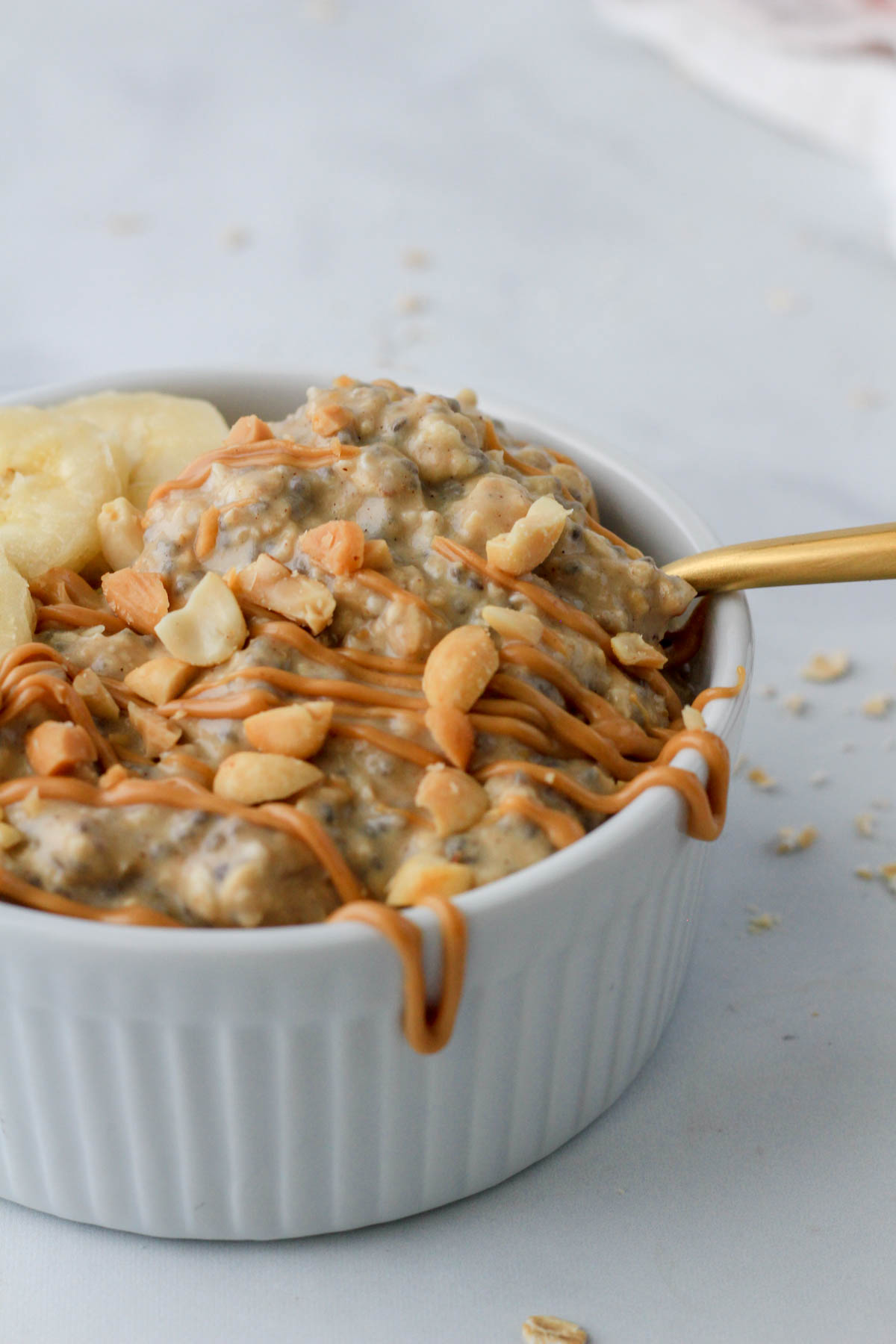 A white bowl with vegan vanilla protein peanut butter overnight oats with peanut butter and peanuts with a gold spoon in the bowl.