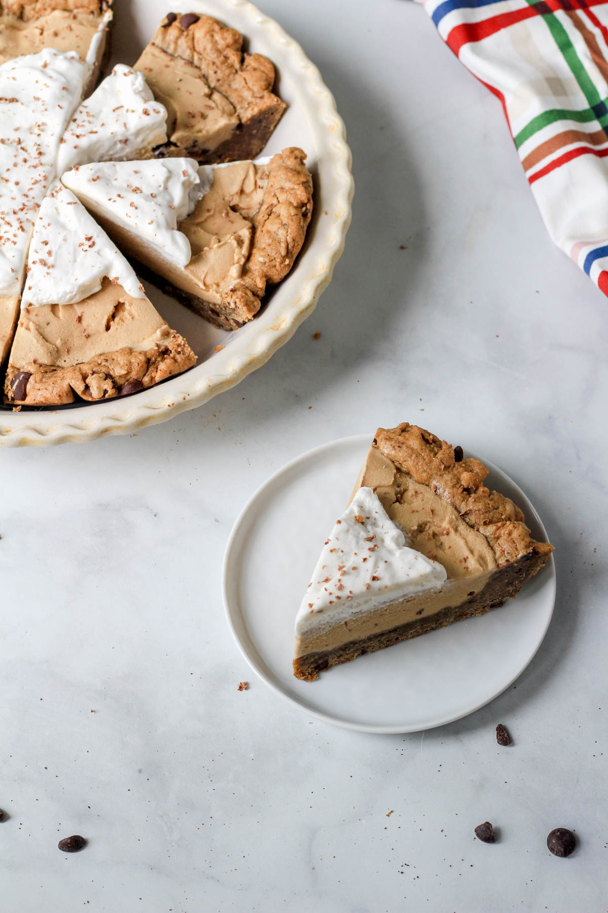 A small white plate with a slice of dairy-free coffee chip ice cream pie topped with dairy-free whipped cream with a pie pan in the back left corner with coffee chip ice cream pie.