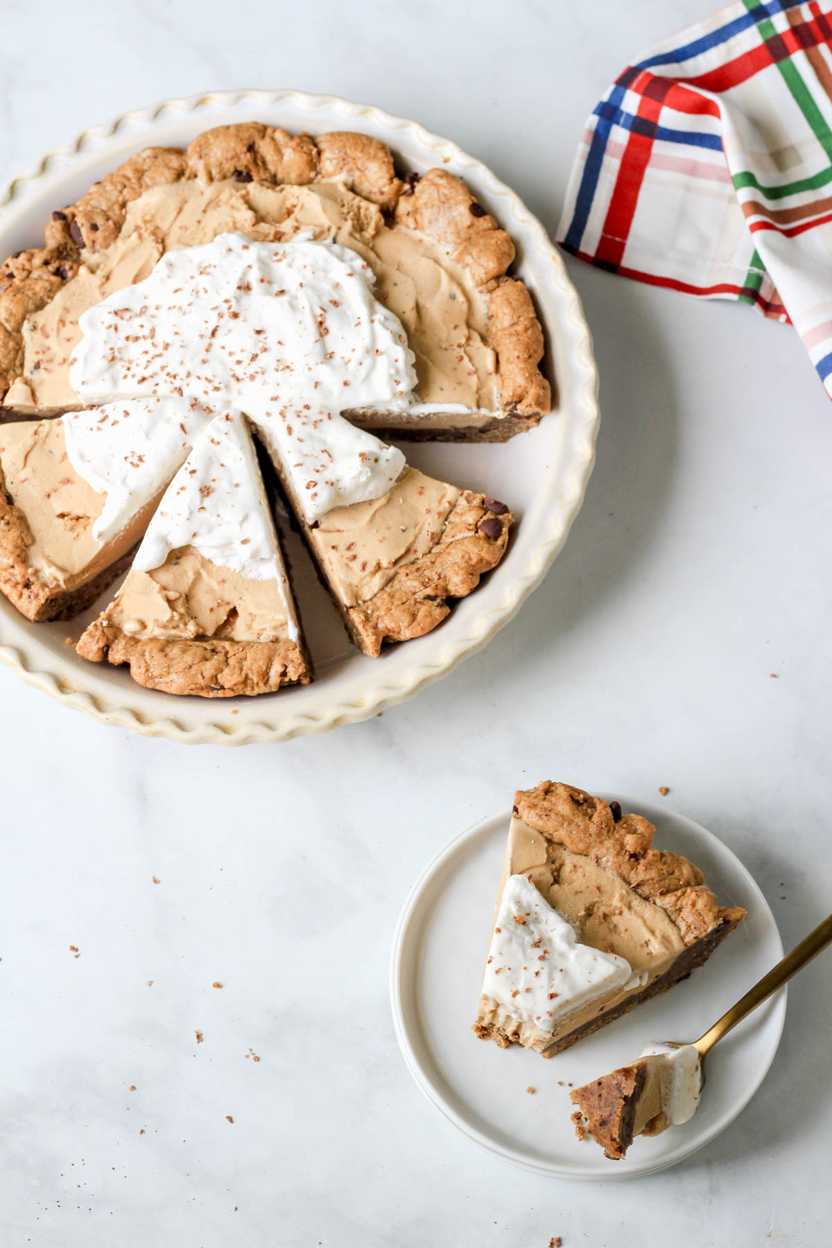 A small white plate with a slice of coffee chip ice cream pie and a fork with a pie pan of coffee chip ice cream pie in the back left corner.