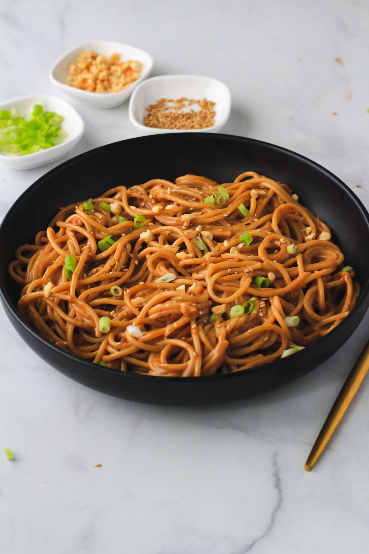 A black bowl filled with 10-minute peanut noodles and three white pinch bowls in the back with toppings.