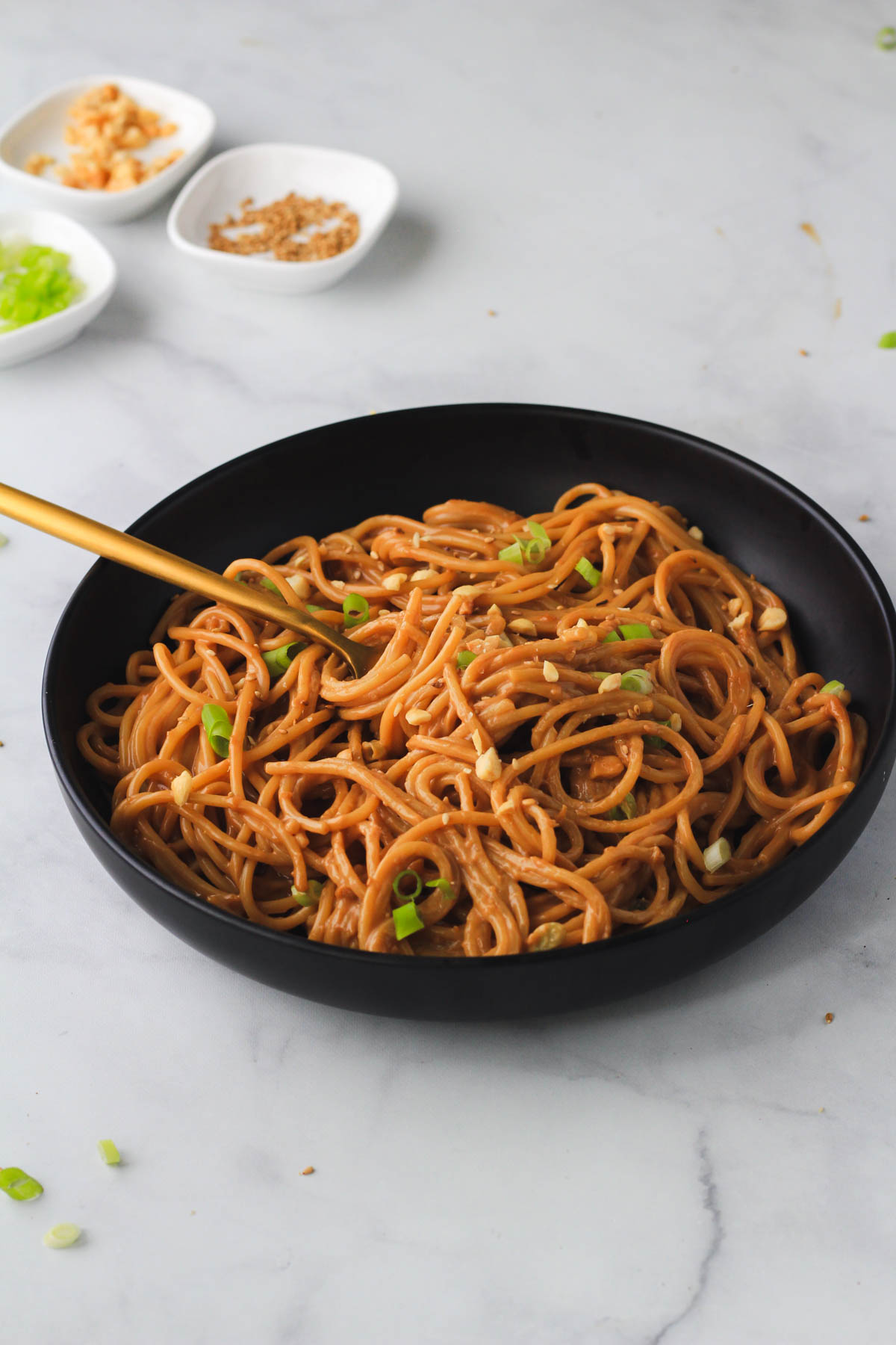 A gold fork in a black bowl of easy 10-minute peanut noodles topped with green onion, sesame seeds, and chopped peanuts.