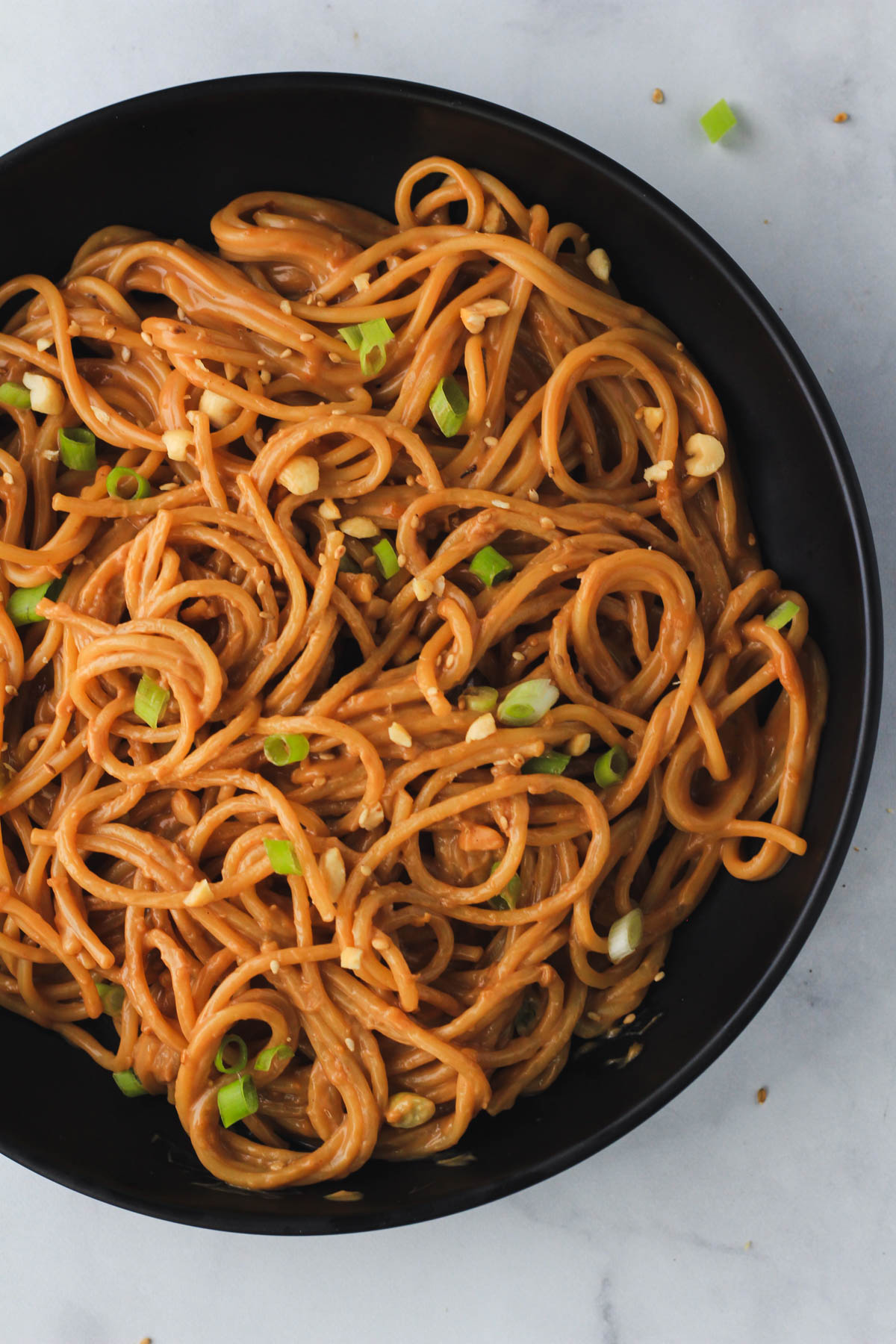 A top down picture of a black bowl of easy, creamy peanut noodles topped with green onion, sesame seed, and chopped peanuts.