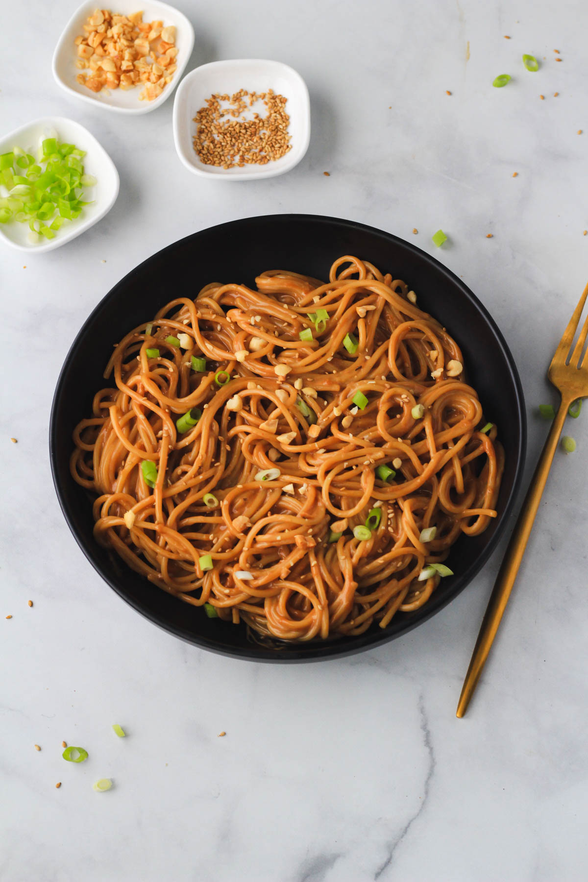 A black bowl with creamy peanut noodles topped with green onion, sesame seeds, and salted peanuts on a grey counter with pinch bowls in the top left.