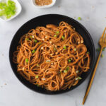 A black bowl of creamy peanut noodles topped with green onion and a gold fork to the right of the bowl.