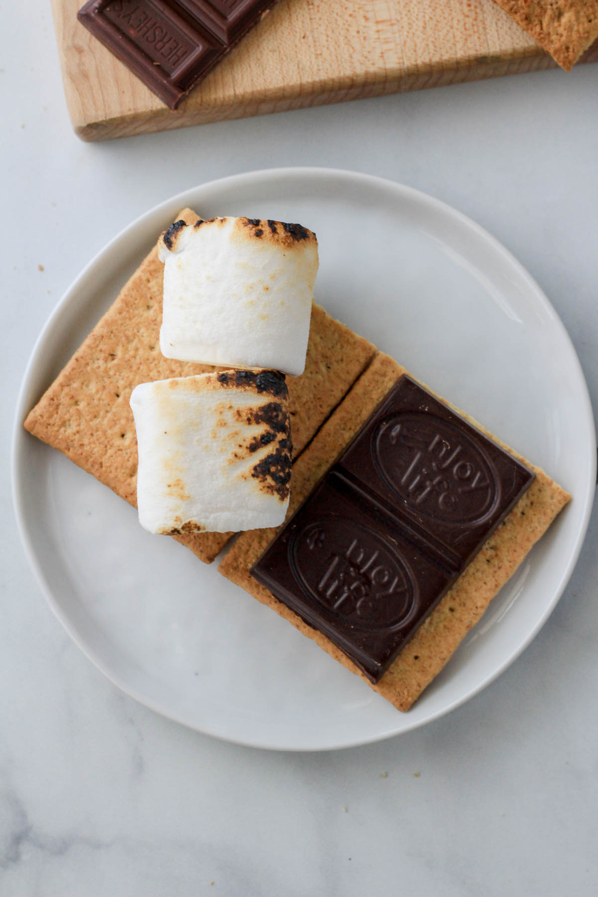 A small white plate with a graham cracker half topped with roasted large marshmallows and a square of dairy-free chocolate.