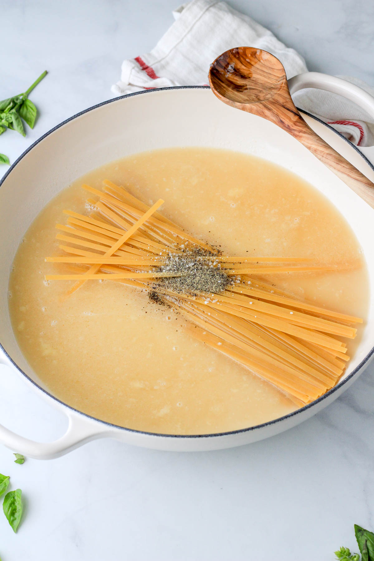 A white pot with pasta, seasoning, and liquids before cooking with a wooden spoon on the top right handle.