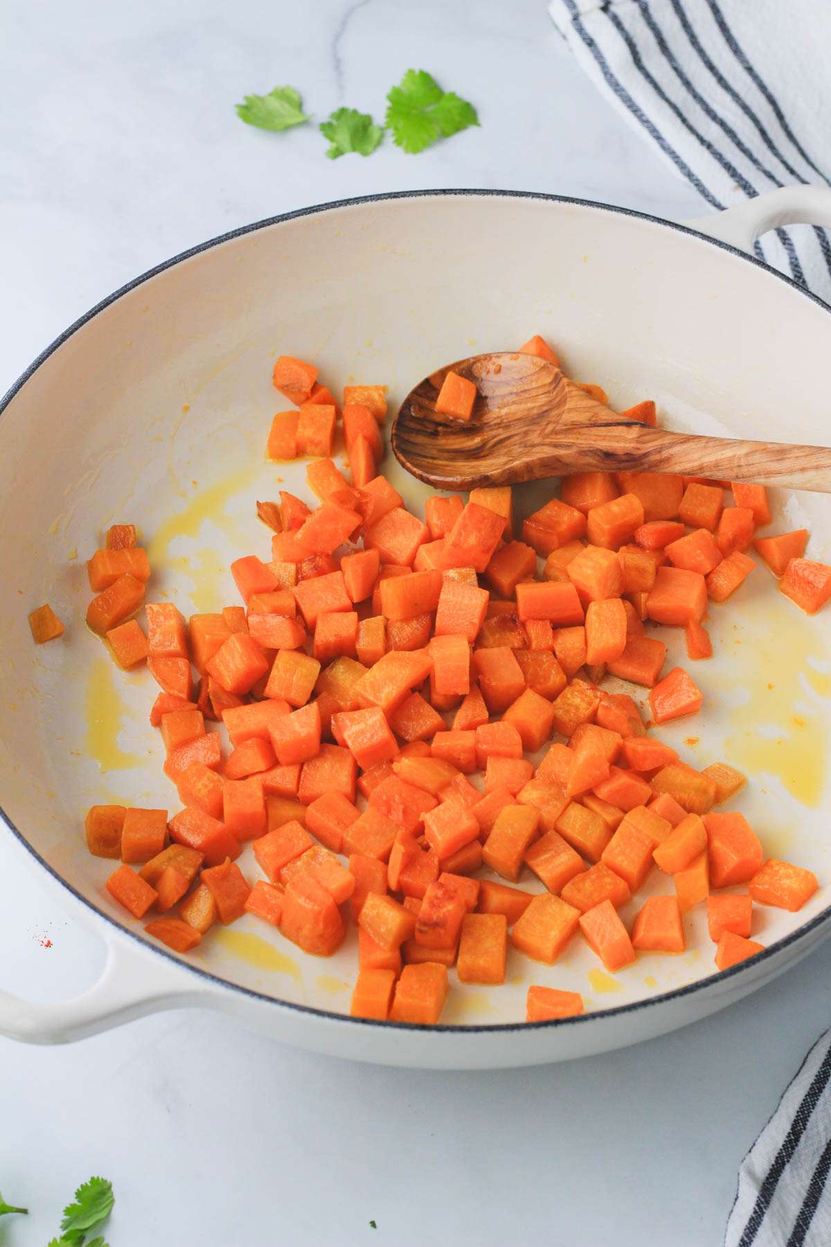 A white skillet with diced sweet potato and a wooden spoon.