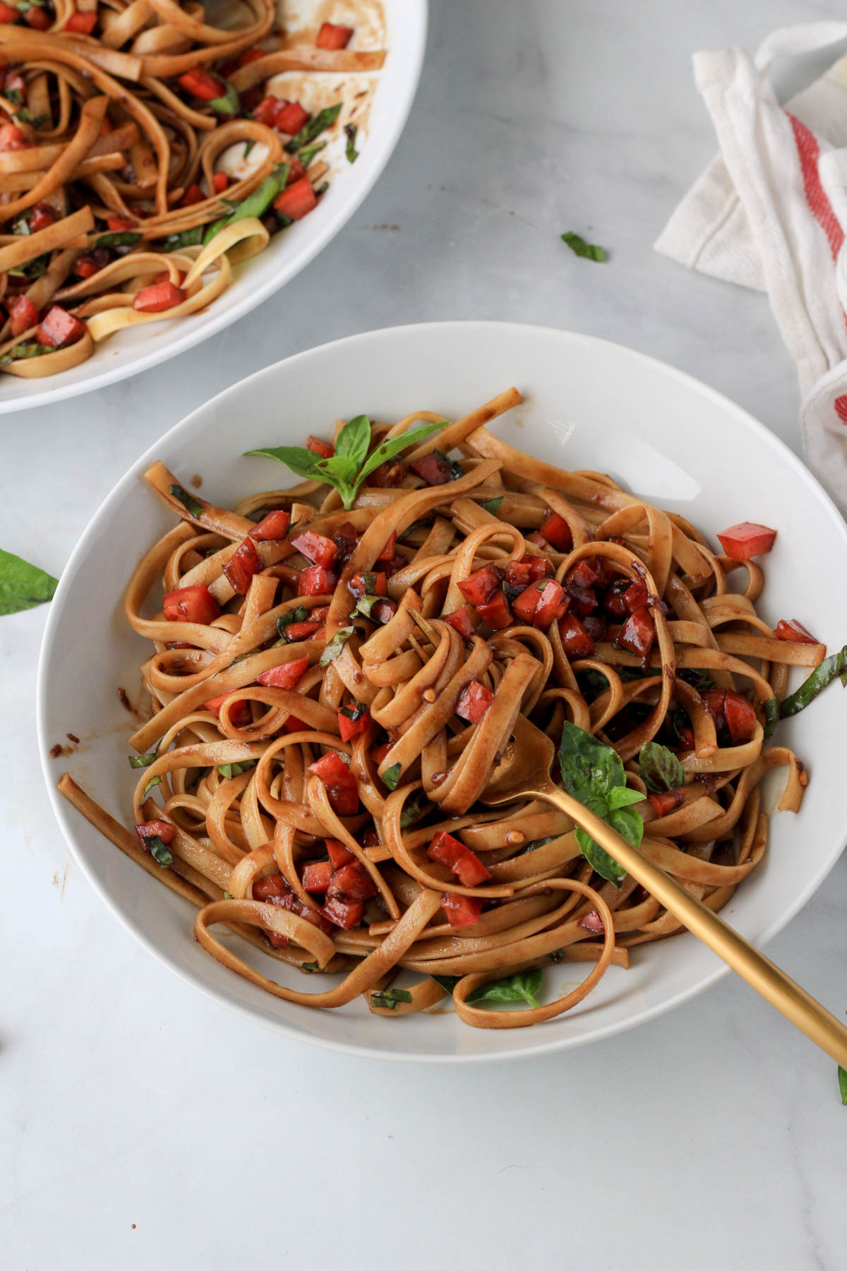 A white bowl of vegan bruschetta pasta and a gold for on a white counter with a larger bowl of bruschetta pasta in the back left corner.