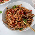A white bowl of vegan bruschetta pasta topped with basil and a gold fork wrapped with vegan pasta.