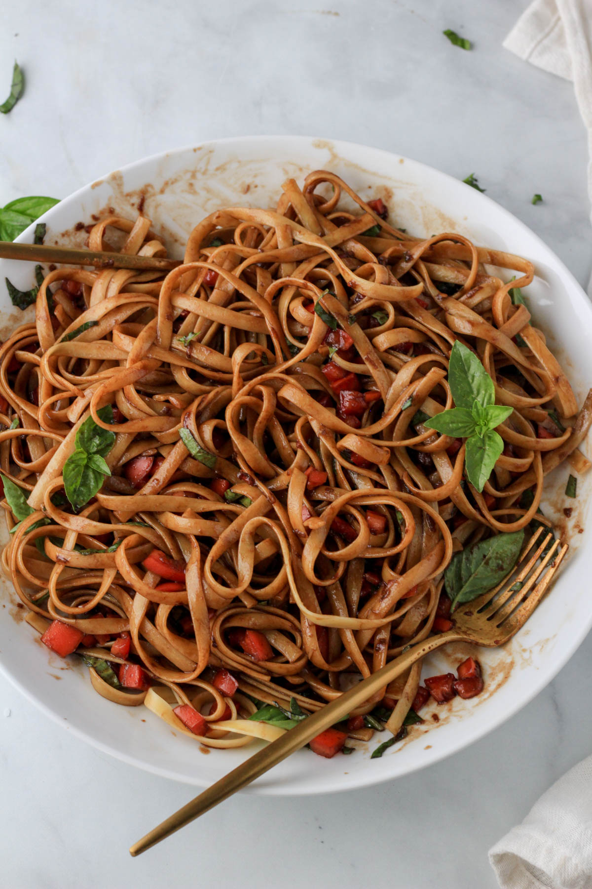 A gold fork and spoon frame a large white bowl of vegan bruschetta pasta topped with basil.