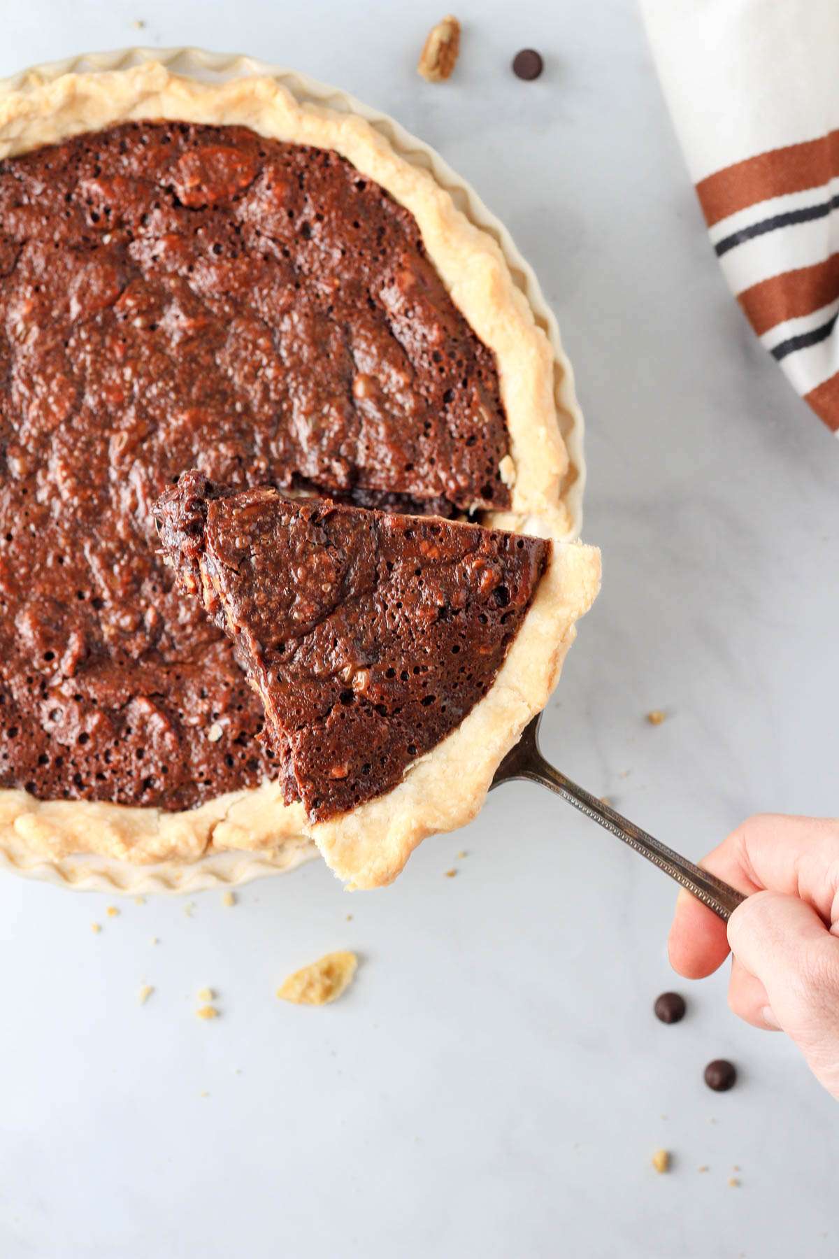 Dairy-free chocolate pecan pie on a pie server with a hand holding the slice above the pie pan.