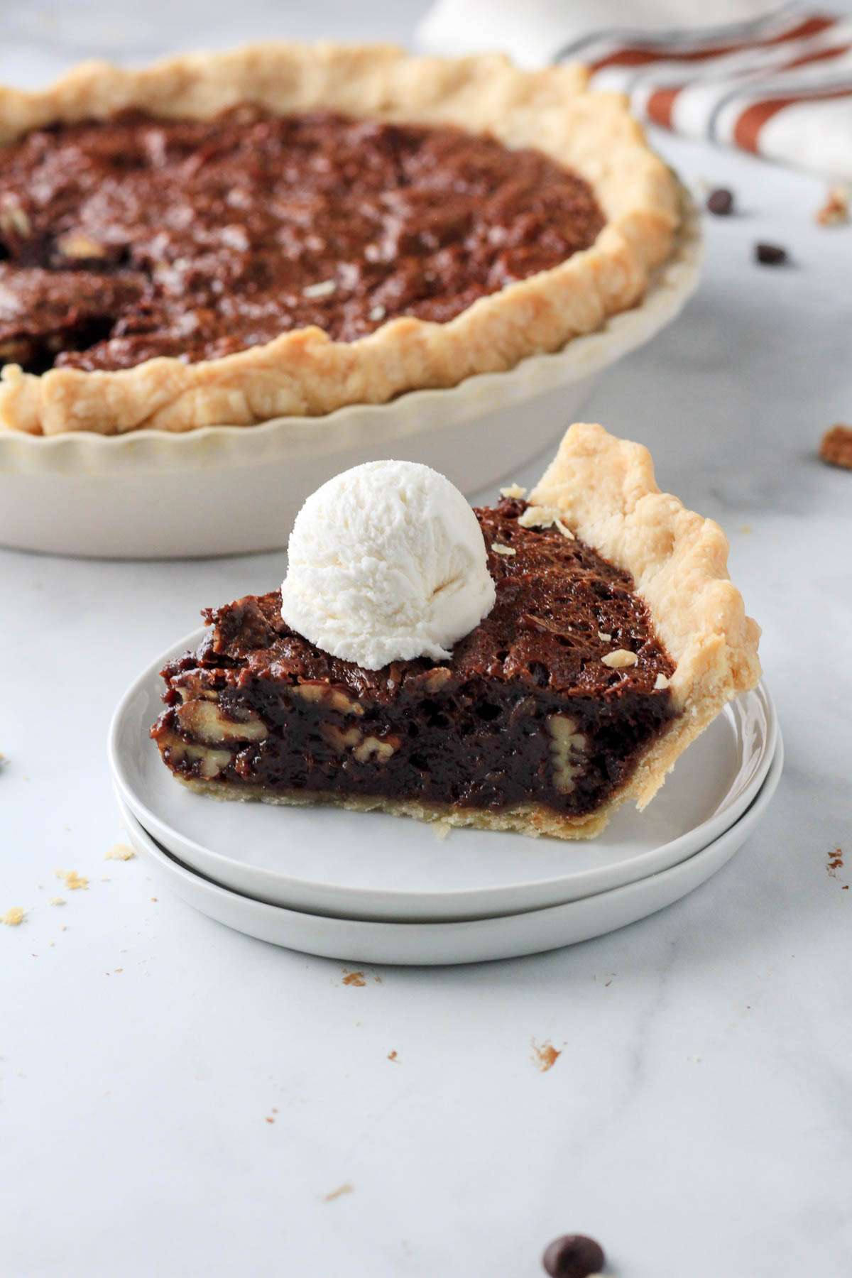 A slice of chocolate pecan pie on a white plate on a white counter in front of the pie pan with the remaining chocolate pecan pie.